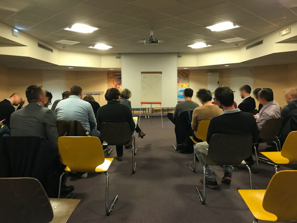 Salle de séminaire vue du fond : une vingtaine de participants assis face à un paperboard, des chaises jaunes vides au premier plan