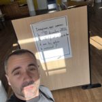 Selfie vertical d’un homme devant un panneau d’atelier collectif, affiche manuscrite « Imaginons, nous sommes le 30 décembre 2026, le collectif est au top du top, à quoi les autres le verraient ? », salle de réunion aux murs jaunes, tables et chaises en arrière-plan, parquet en bois, colonnes blanches, lumière directe du soleil, ombres géométriques marquées, cadrage smartphone en légère contre-plongée, format portrait 3:4, photographie documentaire naturelle, netteté homogène, contraste lumineux élevé