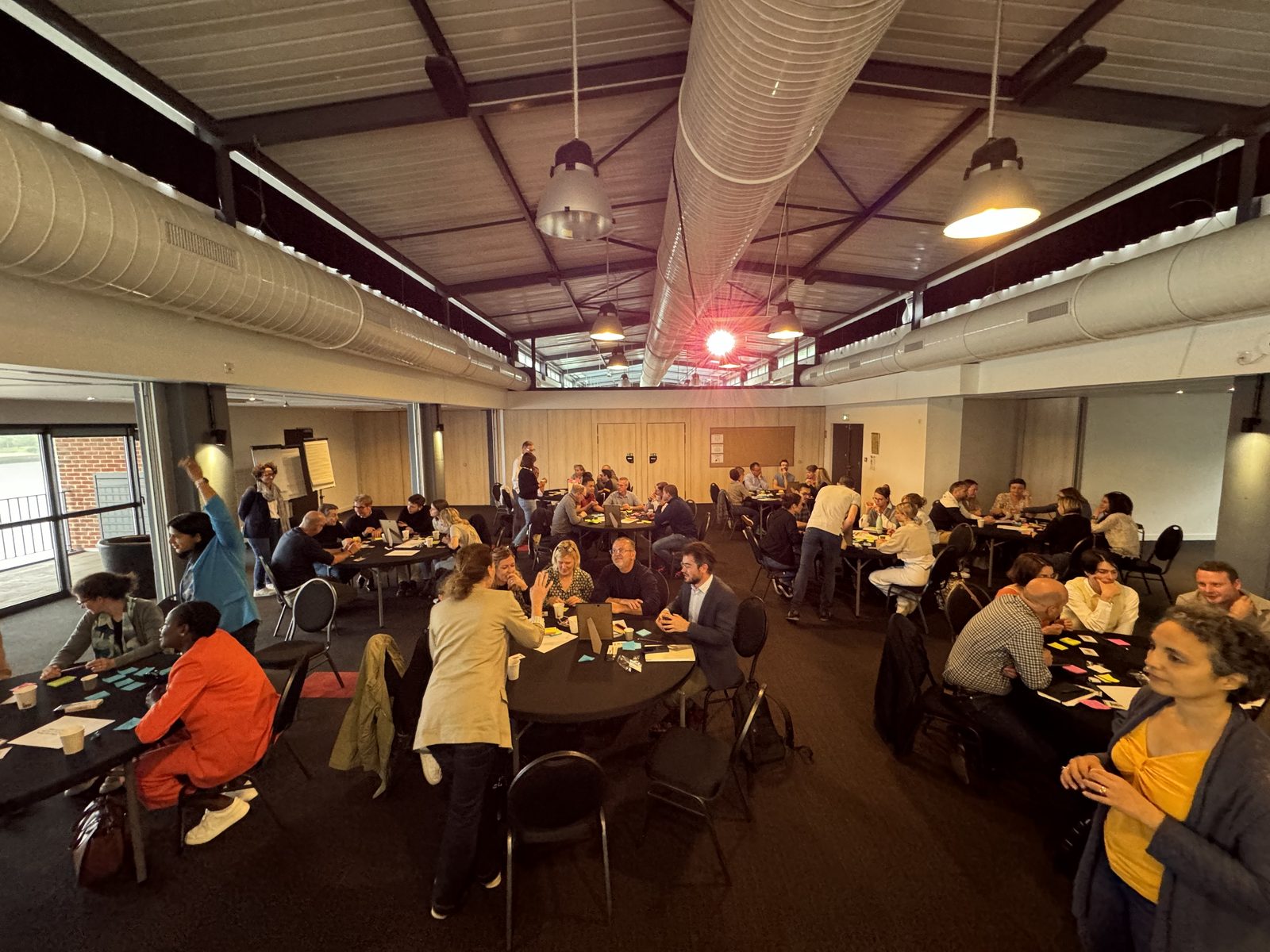 Grande salle de séminaire en plein travail, une vingtaine de tables rondes et des conversations en même temps